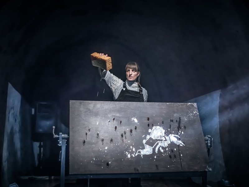 Eine Frau steht hinter einer schwarzen Tafel, auf der ein kleines Mädchen mit Kreide gezeichnet wurde. Der Schwamm, den sie über die Tafel hinausstreckt, tropft mit Wasser.