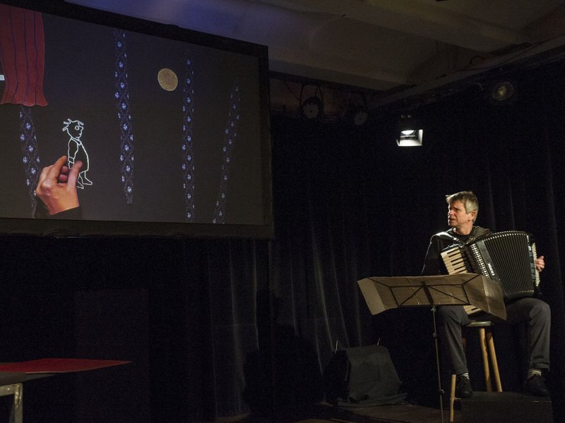 A screen onto which videos of figures made of pieces of paper are projected. A man sits diagonally in front of the screen and plays the accordion while looking at the screen.