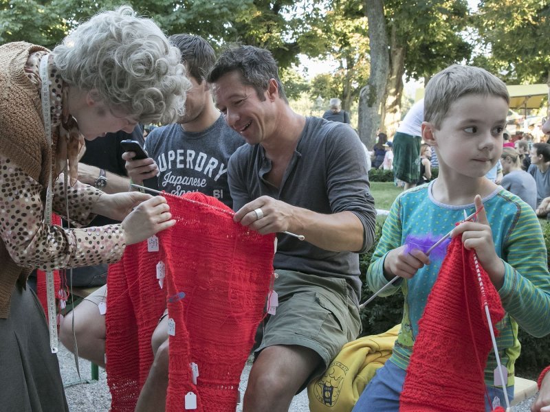 A woman is helping a man to knit a red scarf. Around them, additional people are knitting red scarves, for example a little boy on the right.