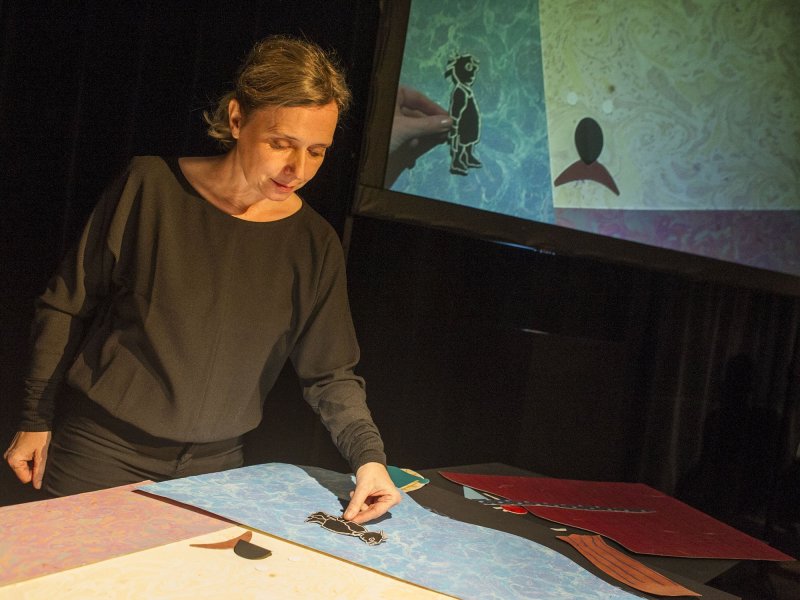 Photo of a woman moving figures made of pieces of paper on a table. The filmed figures are projected onto a screen.