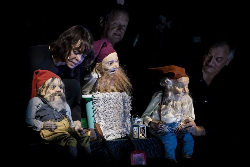 Three people on a dark stage, each holding a bearded puppet wearing a red, pointed cap. Multiple items, like a bucket, a cleaning rag and a latern are propped up on a table between them.