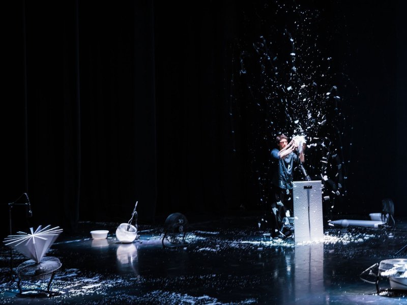 A stage with a few items of daily use and a lot of styrofoam crumbs. A standing man rips more styrofoam with his hands.