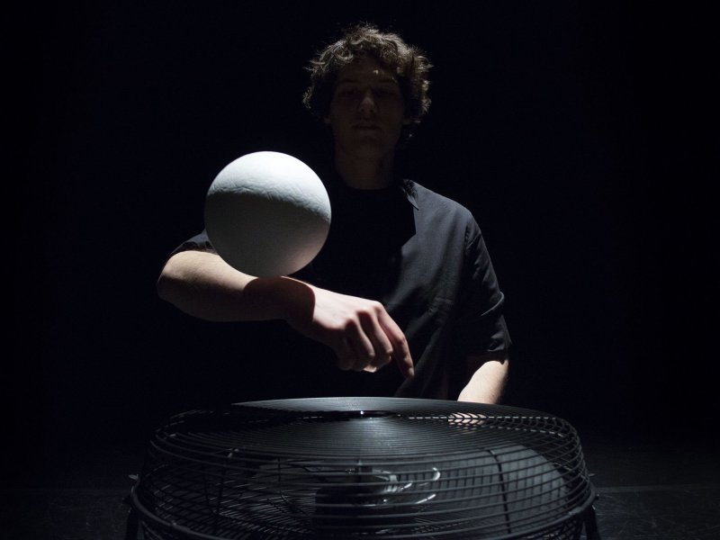 A man sits on the floor of a stage and levitates a large white ball over a lying fan.
