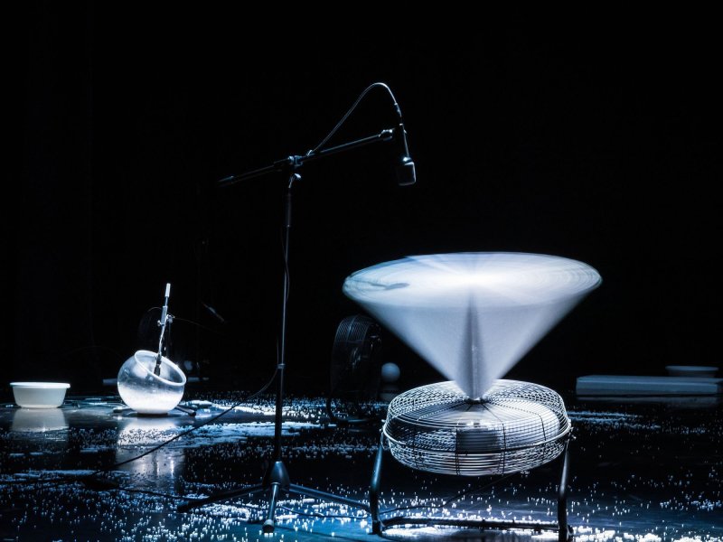 Close-up of a stage with a fan pointing upwards, levitating a white spinning object. There is a lot of styrofoam crumbs in the background.