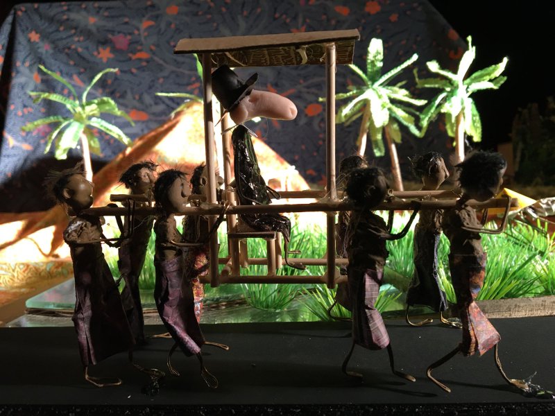 Close-up of crafted, abstract black human figures carrying a sedan chair with a white puppet.