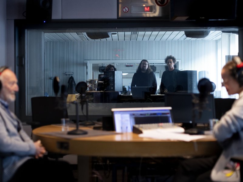 Zwei Personen mit Kopfhörern sitzen sich in einem Tonstudio an einem Tisch gegenüber. Im Hintergrund stehen drei Personen in einem Nebenraum mit Glaswand, die die Personen am Tisch betrachten.