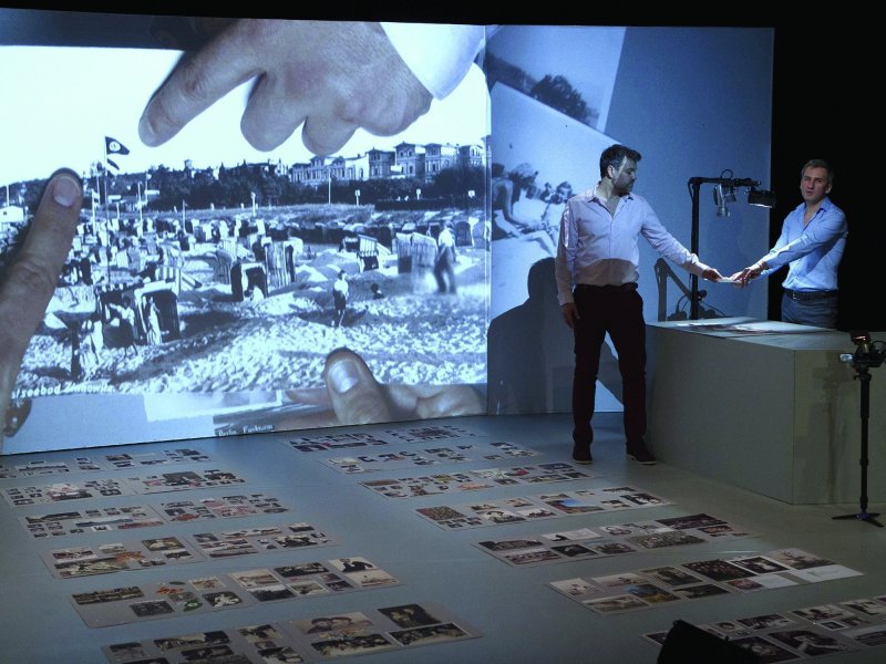 Eine Bühne mit einer Leinwand und Papierbögen mit Fotos auf dem Boden. Auf die Leinwand wird ein Strandfoto projiziert, daneben stehen zwei Männer, die ein Foto unter eine Kamera halten.