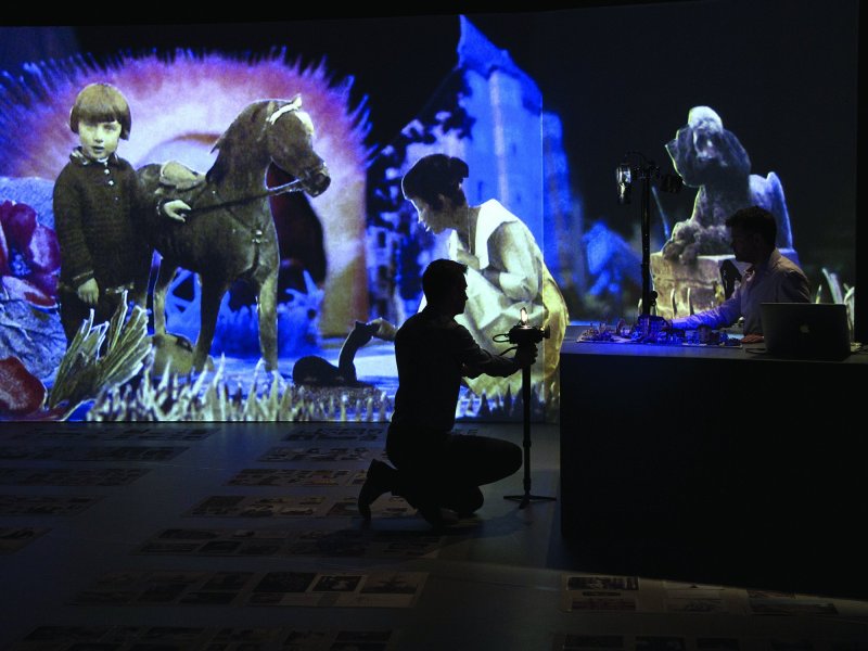 Eine Bühne mit einer Leinwand, auf die der Papierschnitt eines Kindes und eines Ponys projiziert werden. Im Vordergrund kniet ein Mann, der kleine Papierschnitte auf einem Tisch abfilmt.