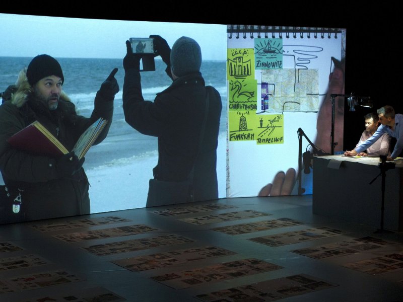 Eine Bühne mit einer Leinwand und Papierbögen mit Fotos auf dem Boden. Auf die Leinwand wird ein Foto von zwei sich besprechenden Männern am Strand projiziert, daneben stehen zwei Männer, die unter einer Kamera zeichnen.