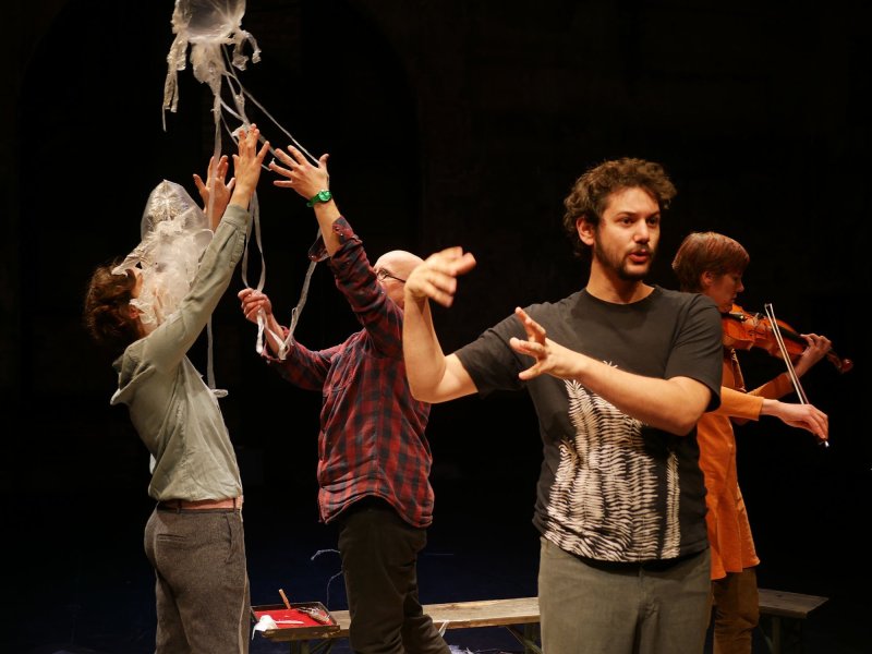 Four people in front of a black background. The one in front is gesturing with his hands, while a woman is playing the violin to his right. The last two are reaching up towards an inflated plastic bag with long, thin plastic strips hanging from it.