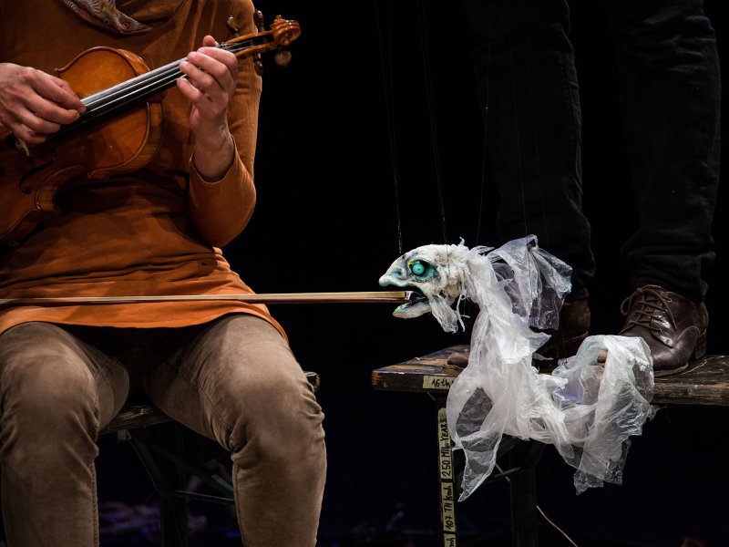 Cutout of a person playing the violin with only her hands. A fish-like creature floating next to her is holding the violin bow in its mouth. Behind it, there is a person standing on a wooden stool.