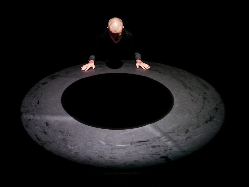 A man is leaning towards the ground while supporting himself with his two hands. His lower half is disappearing into shadows. There is a large black hole in the ground in front of him.