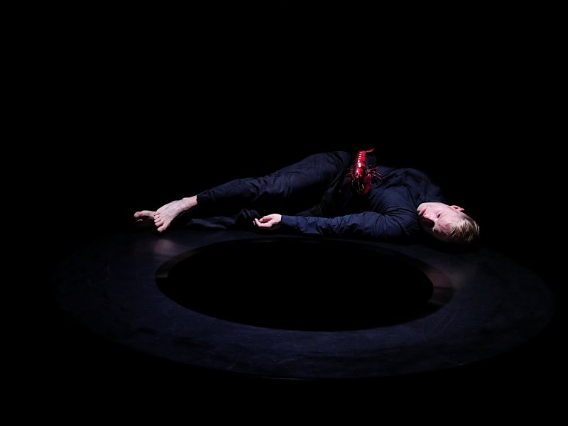 A man dressed in black is lying sideways on the floor of a dark stage, with his upper body is facing upwards. A red lobster is placed on his stomach. In front of him, there is a large black hole in the floor.