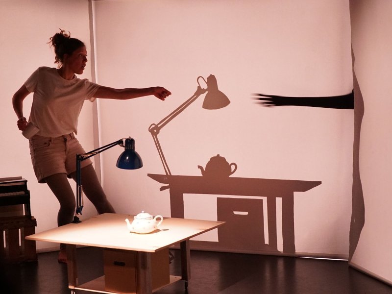 A woman dressed in white is holding a cup in her right hand and reaching her left arm forward. Her arm, as well as the table with teapot and desk lamp placed in front of her, cast a shadow on the wall behind her.