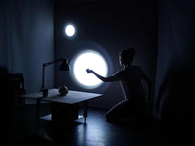 A woman is kneeling on the floor of a dark room. Two points of light can be seen on a white wall behind her. In front of her is a small table with a desk lamp, a cup and a teapot.