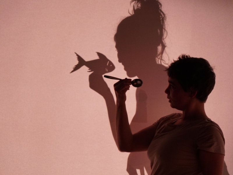 Close-up of a woman holding a spoon up to her face. The shadow cast behind her is imitating that pose, but holding a fish instead of a spoon.
