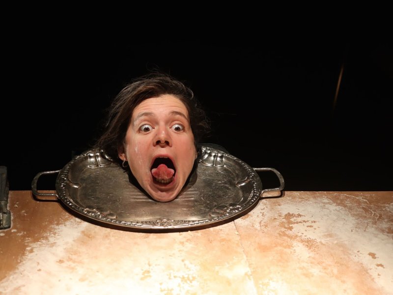 The head of a screaming woman sticking out her tongue lies on a silver tray placed on a table full of flour.