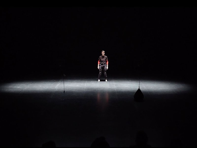 Photo of an empty stage with a young man standing upright in the center.