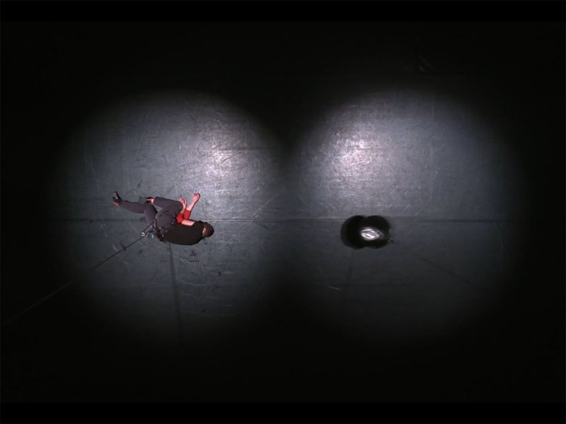 Zwei Rampenlichter auf einer schwarzen Bühne. In einem liegt ein Mann mit Klettergurten. In dem anderen schwebt ein schwarzes Gewicht. Beide sind über ein Seil an der Decke befestigt.