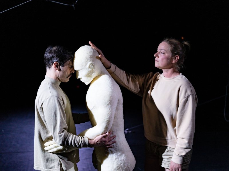 Ein Mann und eine gleich große, weiße Puppe halten sich gegenseitig an der Taille und legen ihre Stirnen aneinander. Hinter der Puppe steht eine Frau, die den Kopf der Puppe gegen den Kopf des Mannes drückt.