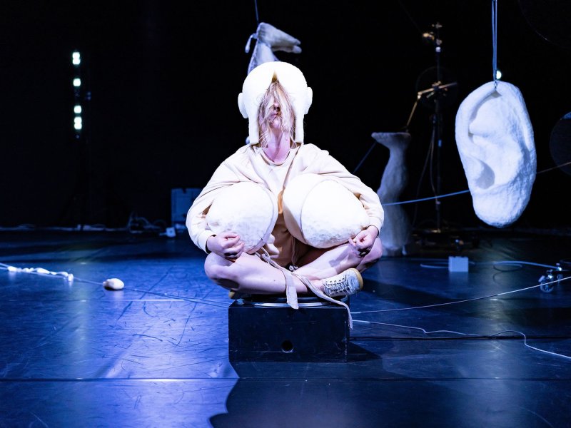 A woman sitting cross-legged on a stage, holding two huge papier-mâché breasts to her body.