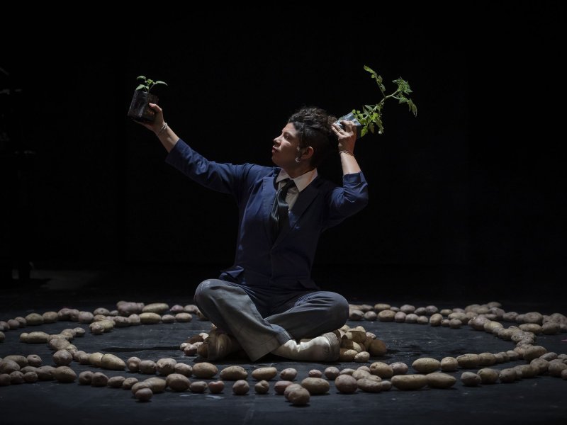 A woman is sitting cross-legged on the floor in front of a black background and is surrounded by potatoes. She’s holding two plants in her hands, stretches them up in the air and looks after them.