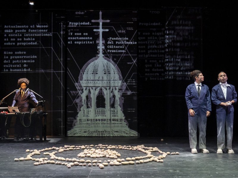 A stage with three people wearing suits. Two are standing next to each other toward the right edge of the image, while the third person is standing behind a desk to the left. Behind them, a schematic of a building is projected. In the center of the stage, some potatoes are arranged in a circle.
