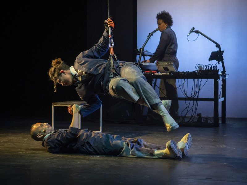 A woman wearing climbing gear is let down from the ceiling, hanging half a meter above the floor. A man is lying below her. They’re clasping their hands together while looking at each other.