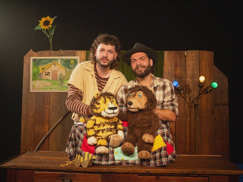 Zwei junge Männer stehen nebeneinander, vor Ihnen zwei Stofftier-Puppen, einTiger und ein Bär. Im Hintergrund eine Holzwand, eine Lampe mit farbigen Birnen, ein Bild und eine Sonneblume.