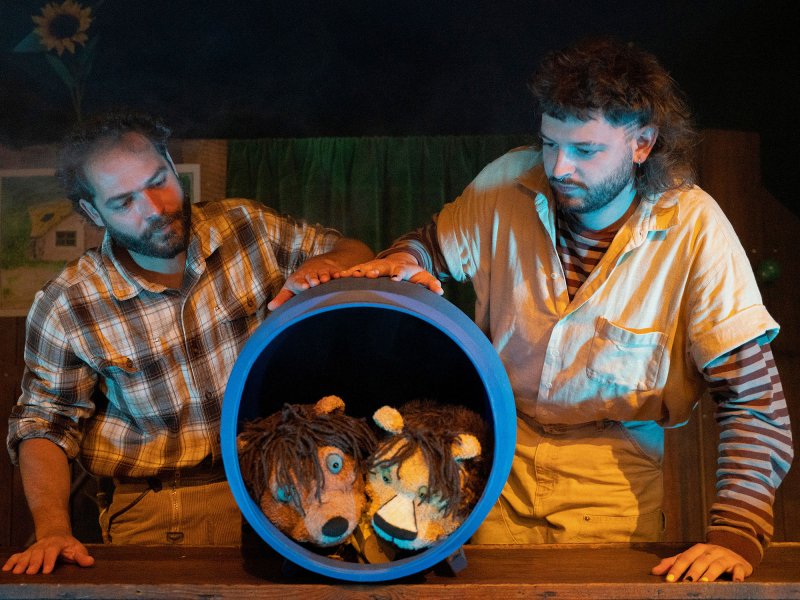 Two stuffed animal dolls, a lion and a bear, are peeking out of a blue plastic tub, which is being rolled and watched by two young men.