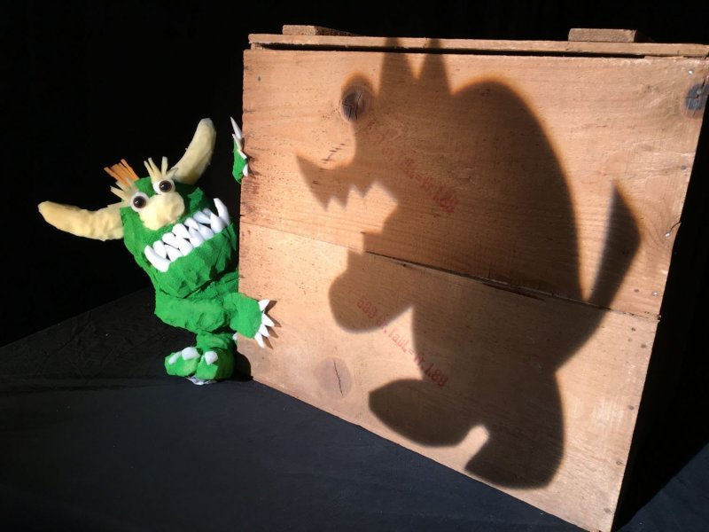A green monster looking scared a tthe shadow of another bigger monster, which is projected onto a wooden box.