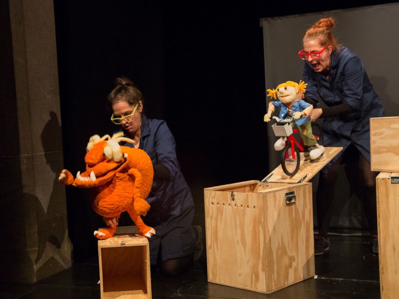 Two women playing rag dolls on wooden boxes. The woman on the left controls a brown monster, the woman on the right a girl riding a bicycle.