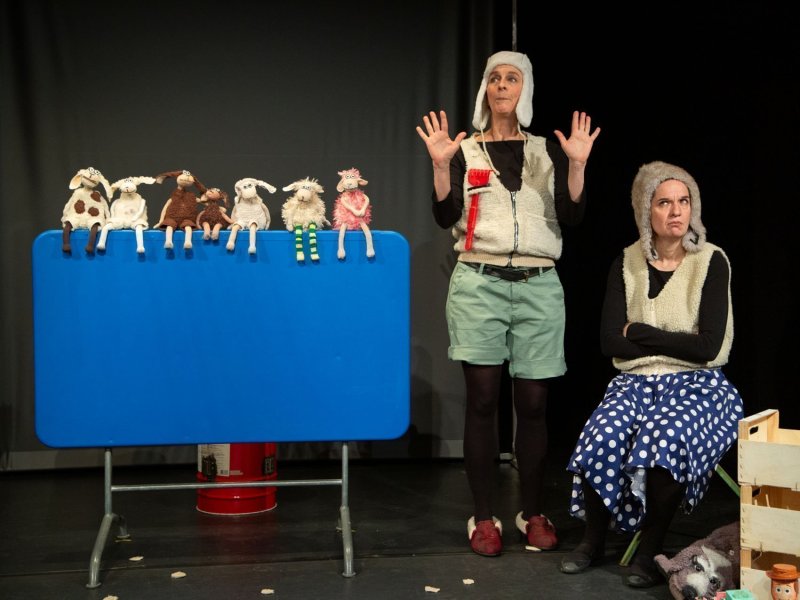 Eine Bühne mit einem Klapptisch mit senkrechter Platte, auf dessen Kante einige kleine Schaf-Plüschtiere aufgereiht sitzen. Neben dem Tisch sitzen bzw. stehen zwei Frauen in Schafspelzkostümen.