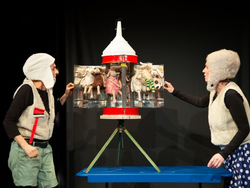 A small self- built, unfolded rocket with sheep inside. The flaps are held by two women in sheepskin costumes, that look worried