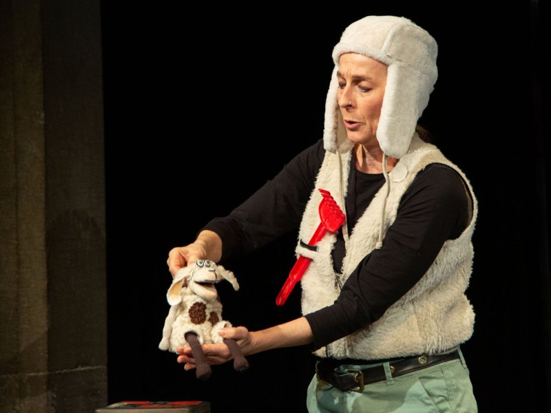 A woman in a sheepskin costume holding and controling a small plush sheep toy.