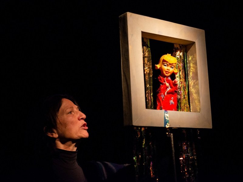 Photo of a feminine hand puppet in a frame with glitter stripes. She is controlled by a puppeteer.
