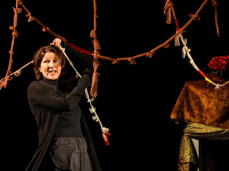 A puppeteer enthusiastically tugs at a multicolored garland made of condoms hanging around her.