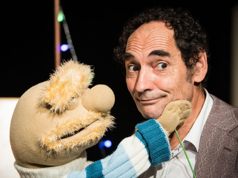 A man with short curly hair looks expectantly to the side while a small rag doll with a beard and bushy eyebrows places it‘s right hand on the man's cheek.
