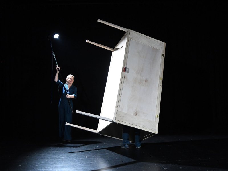 Zwei Personen in blauen Overalls stehen auf einer dunklen Bühne. Die Linke hält einen Scheinwerfer an einem langen Stab auf die Rechte, die in einer großen Holzbox mit Tischbeinen steht.