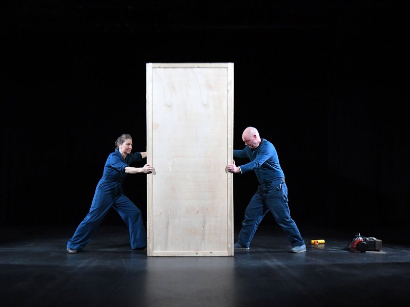 Two people in blue overalls are holding onto a big wooden box from each side.