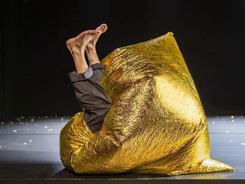 Eine Frau liegt auf einer dunklen Bühne. Ihr Oberkörper ist komplett in einen Sack aus goldener Folie versunken, während sie ihre Beine nach oben streckt.