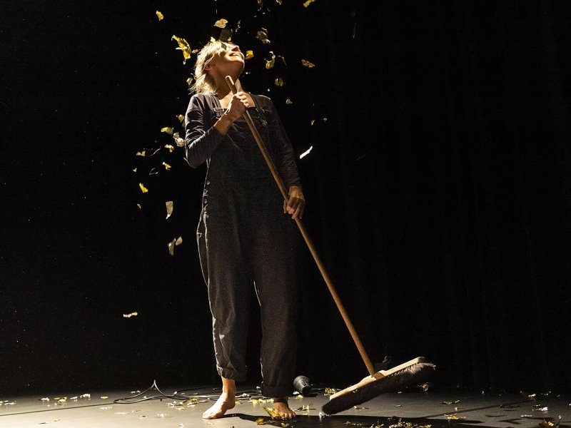 A woman with a broom in her hand is standing on a dark stage, while looking upwards delightedly. Pieces of golden foil fly around her.