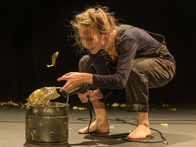 A woman with disheveled hair stands bent over on a stage, while directing a hairdryer at some gold foil that is inside of a bucket.