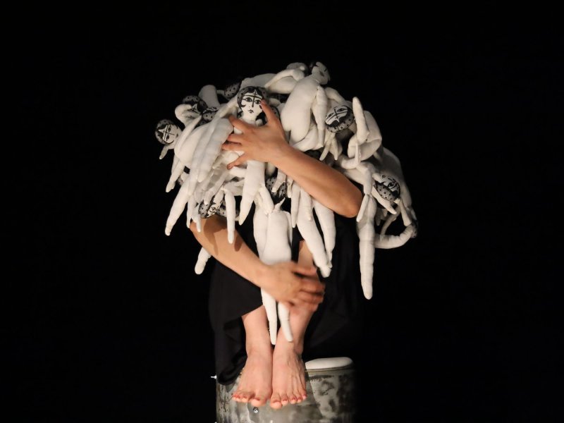 A woman is sitting huddled on a stool. On top of her lie many, small, white rag dolls with faces