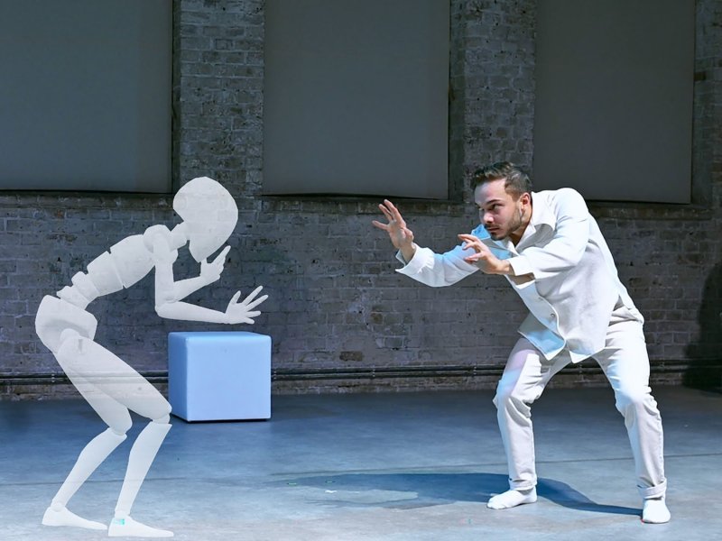 A man in white clothing with a hunched posture is facing a translucent digital figure that imitates his pose.
