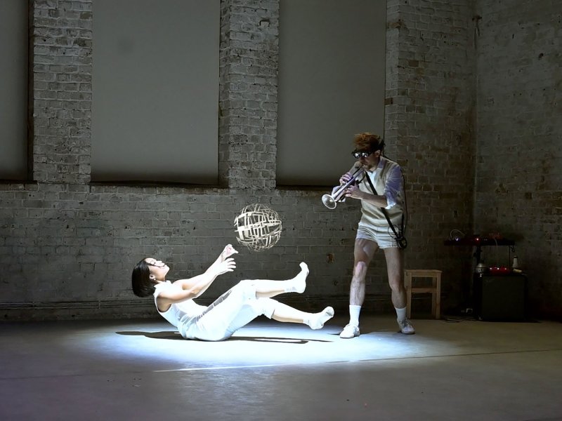 A woman in white clothing is lying on the floor while is stretching her upper body, as well as her arms and legs in the air. Behind her stands a man wearing large black goggles, who plays the trumpet in her direction. Above her legs hover spherically arranged rectangles.