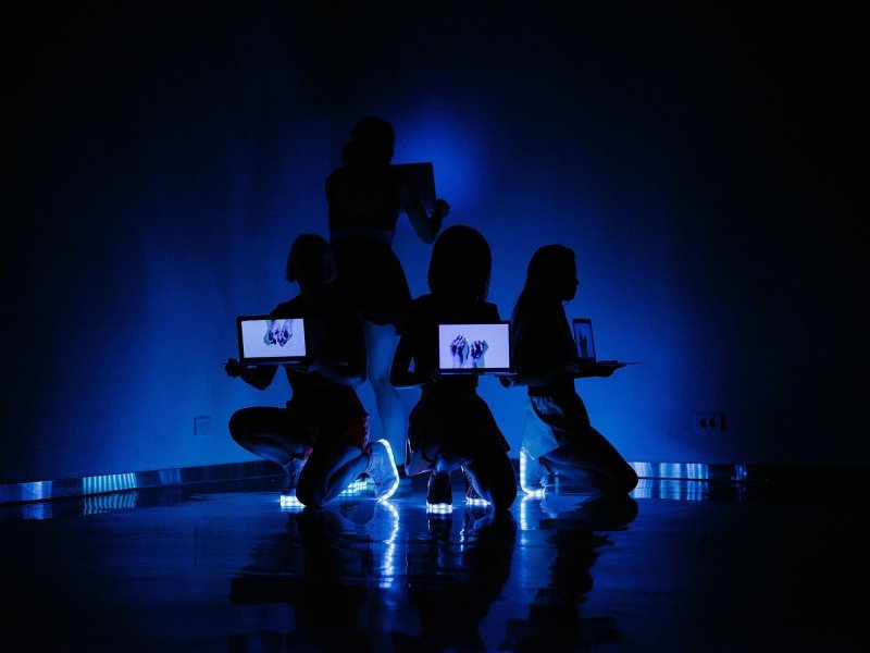 Three women kneel in the corner of a room with another person standing behind them. All of them carry laptops, each showing two hands on the display. The weomen are wearing glowing shoe soles, which are the only source of light illuminating the room.