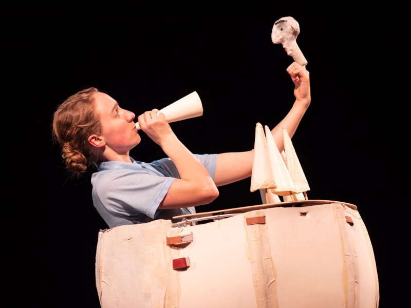 Photo of a young woman dressed as an ice cream seller carrying a cardboard ice cream shop. She talks through a small cardboard cone to a cardboard face stuck on her index finger.