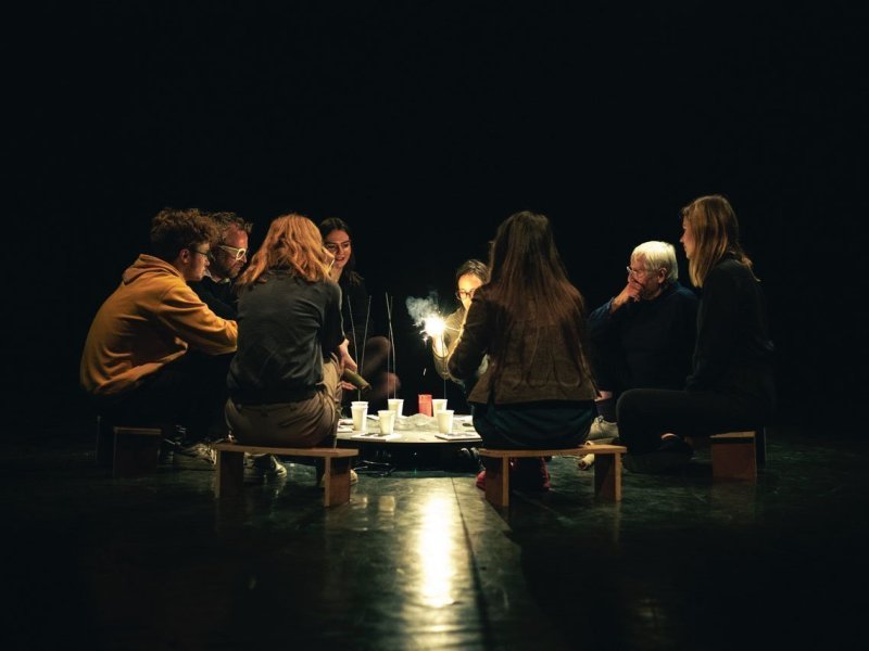 Photograph of a group of people seated around a low table on a stage. One person lights a kind of sparkler, the others watch.