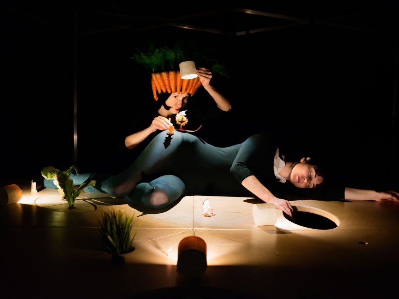 Foto von einer schlafenden Frau auf einem hölzernen Untergrund. Eine zweite Frau hinter ihr lässt eine kleine Figur auf ihrem Körper rutschen und beleuchtet die Szenerie mit einer Lampe.